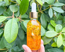 Load image into Gallery viewer, A person holding a 2 oz glass bottle of body oil with a dropper. The bottle is filled with a yellow-hued oil, and it appears to be infused with botanical elements.
